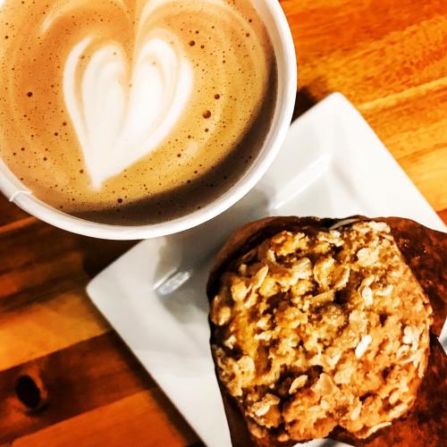 A latte with a heart design on top next to a muffin on a white plate, set on a wooden table.