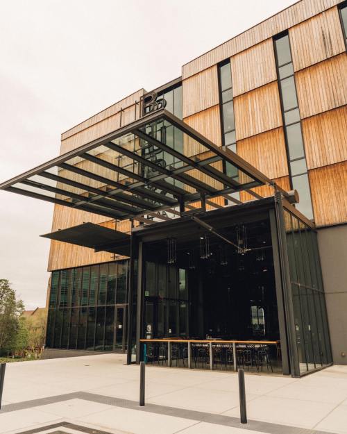 Modern building with wooden facade and large glass entrance, featuring outdoor seating and a sleek design.