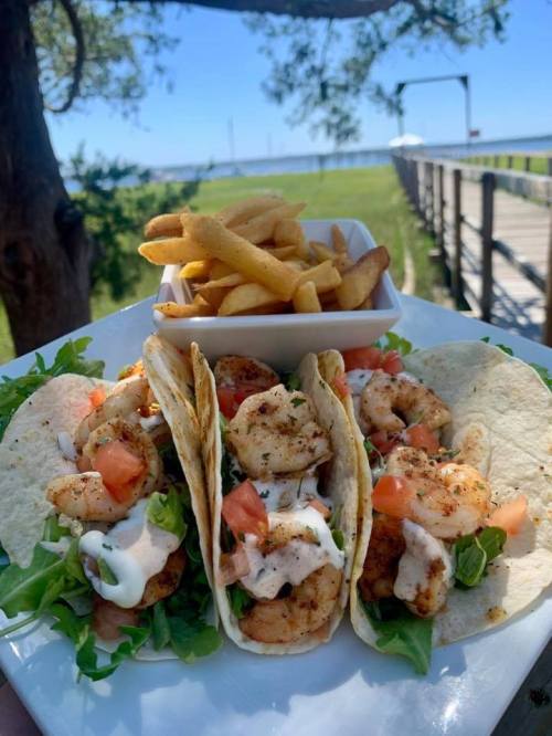 Three shrimp tacos with lettuce, tomatoes, and sauce, served with a side of fries against a scenic outdoor backdrop.