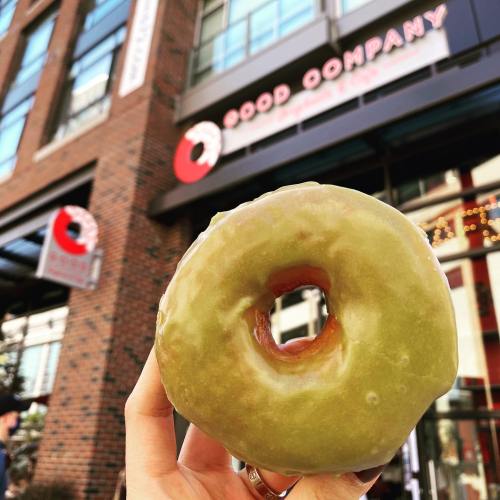 A hand holds a glazed green donut in front of a shop with a sign that reads "Good Company."