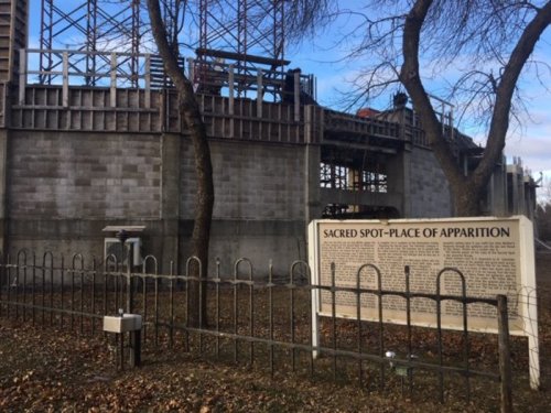 Construction site with a sign reading "Sacred Spot - Place of Apparition" surrounded by a fence and trees.