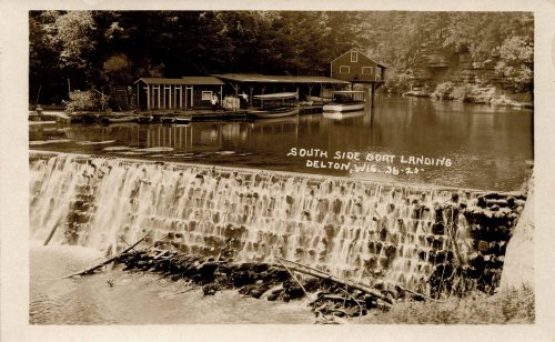 Historic photo of South Side Boat Landing in Delton, WI, featuring a dam and boathouse by a serene waterway.