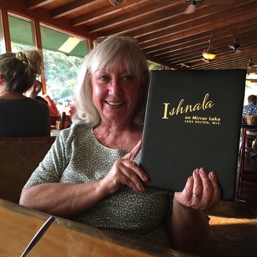 A smiling woman holds a menu at a restaurant, with a cozy interior and diners in the background.