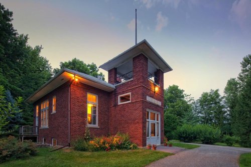 A charming brick house with a tower, surrounded by greenery and illuminated by warm lights at dusk.