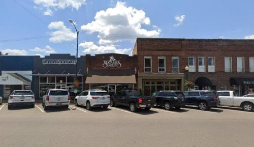 A street view of a small town with shops and parked cars under a blue sky with fluffy clouds.