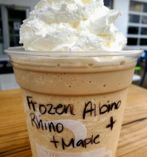 A frosty beverage topped with whipped cream, labeled "Frozen Albino Rhino + Maple" in a clear cup.