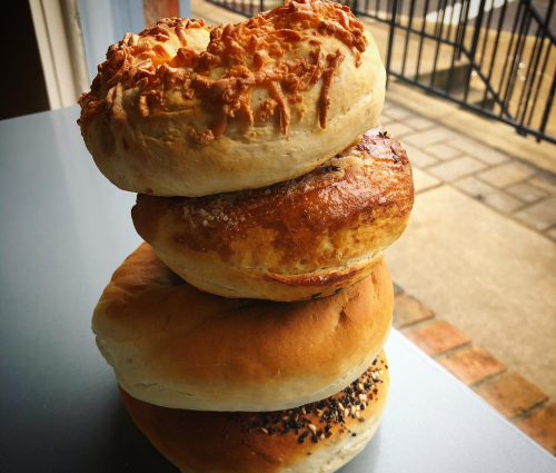 A stack of four freshly baked bagels with various toppings, resting on a gray surface near a window.