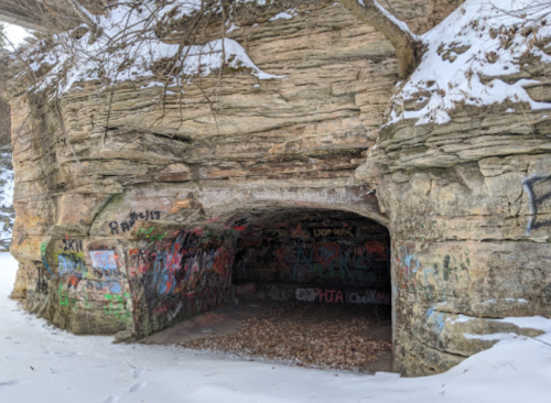 A snow-covered rocky cave entrance with colorful graffiti on the walls, surrounded by bare trees.