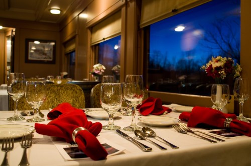 Elegant dining setup on a train, featuring red napkins, glassware, and a view of a twilight sky through large windows.