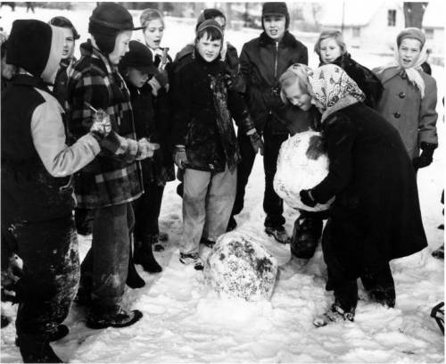 A group of children in winter clothing gathers around two kids building a large snowman in a snowy landscape.