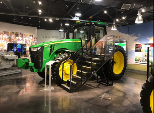 A green and yellow tractor displayed in a museum, with stairs for visitors to access it.