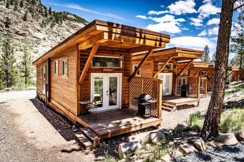 Three wooden cabins with porches, surrounded by trees and mountains under a blue sky with scattered clouds.