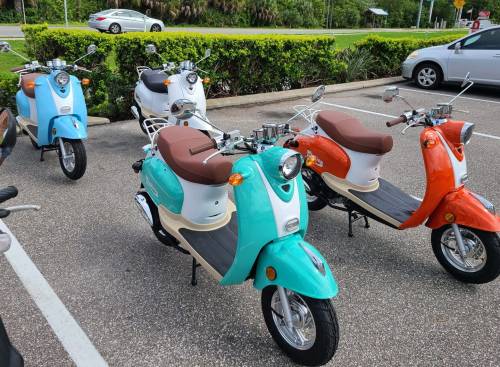 A row of colorful scooters in a parking lot, featuring turquoise, white, and orange models with brown seats.