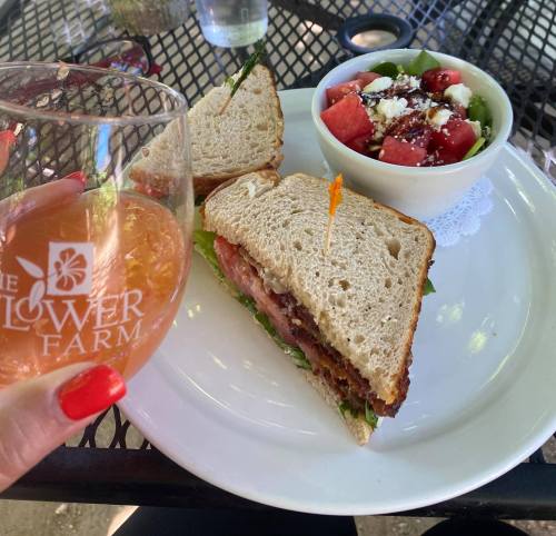 A hand holding a glass of drink next to a sandwich on a plate and a bowl of salad with watermelon and feta.