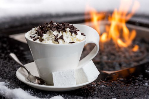 A cozy cup of hot chocolate topped with whipped cream and chocolate shavings, beside a warm fire in a snowy setting.