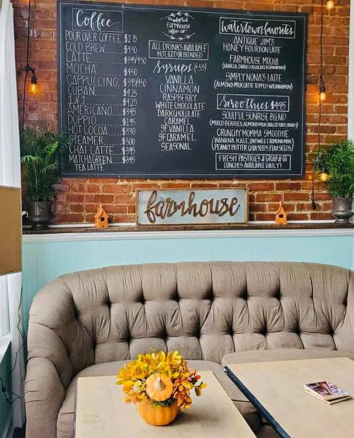 A cozy café interior featuring a chalkboard menu, a tufted sofa, and a small pumpkin centerpiece with flowers.