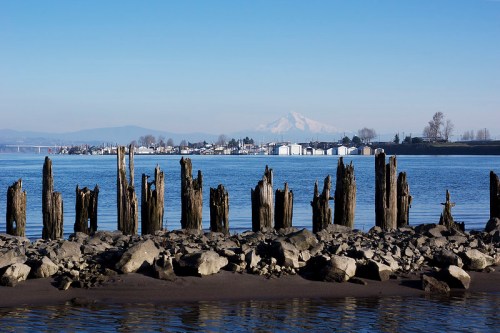 Weathered wooden pilings stand in the foreground, with a calm river and distant mountains under a clear blue sky.