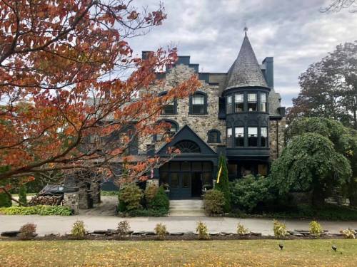A large stone mansion with a turret, surrounded by autumn foliage and a cloudy sky.