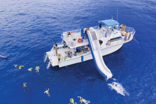 A boat with a water slide is anchored in blue waters, with people swimming and enjoying the sun around it.