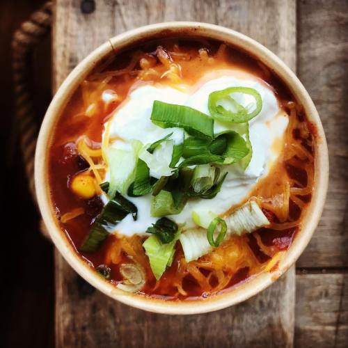 A bowl of chili topped with sour cream, cheese, and green onions, served in a rustic wooden setting.