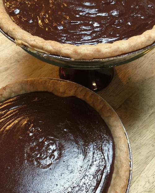 Two chocolate pies in crusts, one on a pedestal and one on a flat surface, showcasing glossy, rich filling.