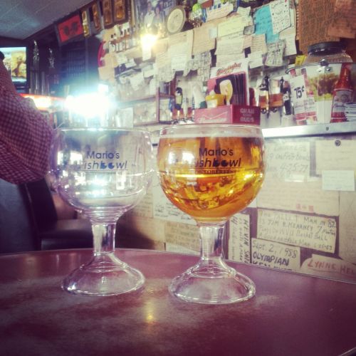 Two large glasses on a table, one empty and one filled with golden liquid, in a bar with a cluttered background.