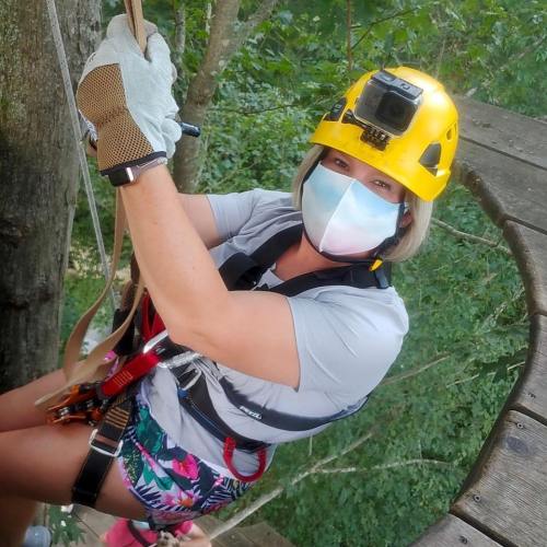 A woman in a yellow helmet and mask is climbing a tree, secured with ropes, surrounded by greenery.