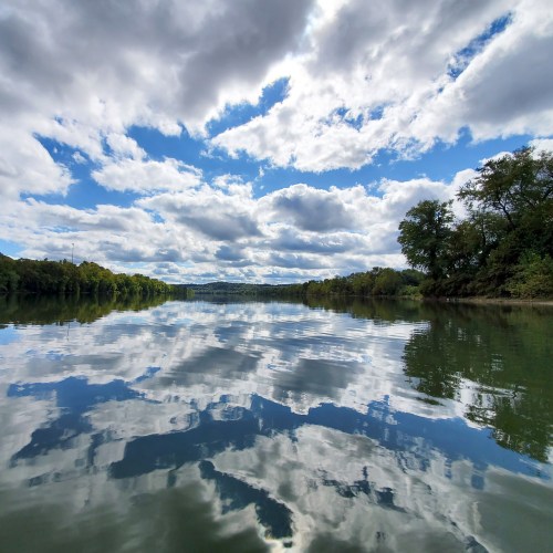 A serene river scene reflecting fluffy clouds and trees under a bright blue sky.