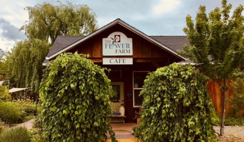 A rustic café building with a sign reading "The Flower Farm Café," surrounded by lush greenery and trees.