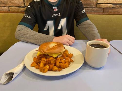 A plate with a sandwich and seasoned potatoes, alongside a cup of coffee, on a table with a person wearing an Eagles jersey.
