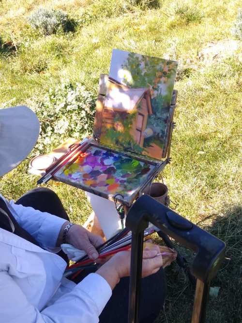 An artist paints outdoors, using a palette filled with vibrant colors and a canvas depicting a rustic scene.