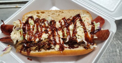 A pulled pork sandwich topped with coleslaw and drizzled with barbecue sauce, served in a foam container.