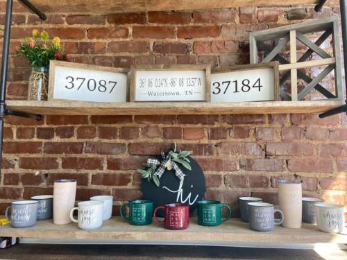 A rustic shelf displays mugs and decorative items against a brick wall, featuring coordinates and a "hi" sign.