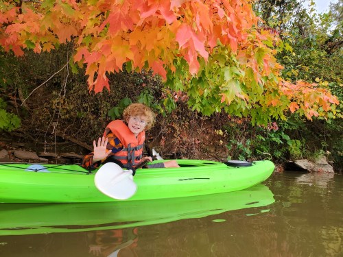 A smiling child in a green kayak waves, surrounded by vibrant autumn leaves reflecting on the water.