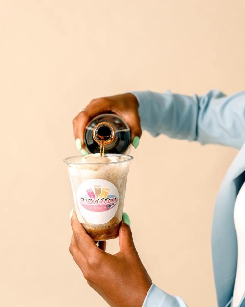 A hand pours soda over a frosty dessert in a clear cup with a logo, set against a light background.