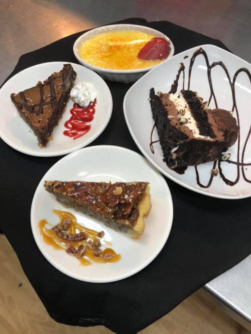 A variety of desserts on plates: chocolate cake, pecan pie, crème brûlée, and a slice of chocolate brownie with toppings.
