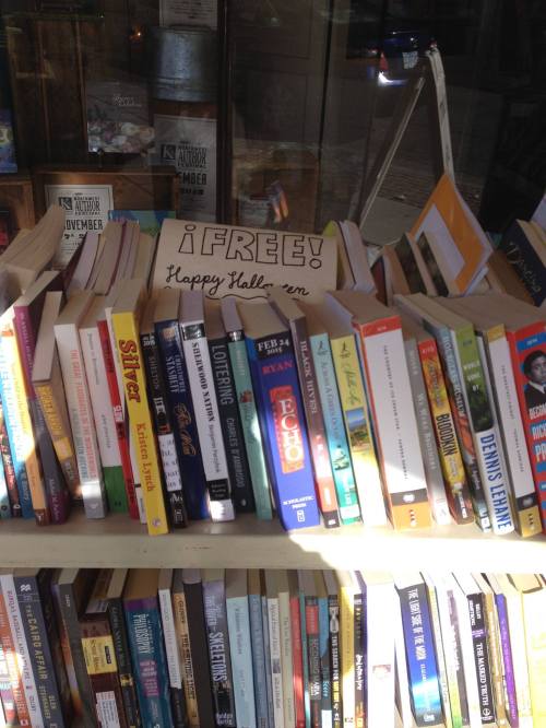 A display of colorful books on a shelf with a sign that says "¡FREE! Happy Halloween!" in a bookstore window.