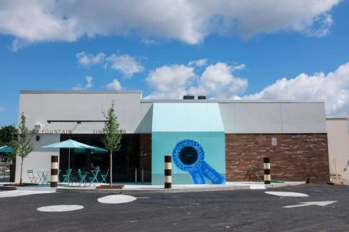 A modern building with a turquoise accent, outdoor seating, and a blue circular feature on the facade under a cloudy sky.