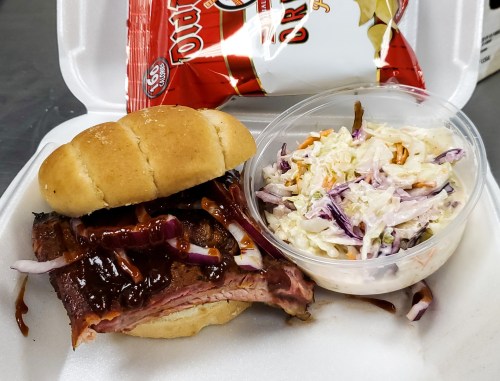 A BBQ sandwich with brisket and onions, served with coleslaw and a bag of chips in a takeout container.