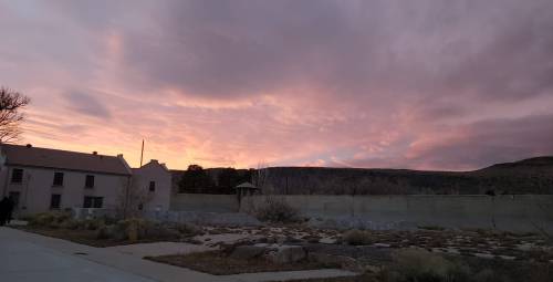 A colorful sunset over a landscape with a building and rocky terrain, framed by clouds and distant hills.
