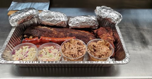 A tray of barbecue featuring ribs, pulled pork, and coleslaw in containers, wrapped items on the side.