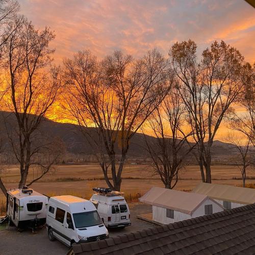 A scenic sunset over a valley, with trees, RVs, and tents in the foreground. Warm orange and yellow hues fill the sky.