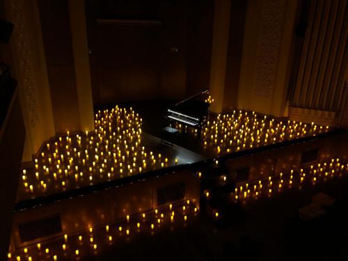 A dimly lit concert hall with a grand piano surrounded by glowing candles, creating a warm, inviting atmosphere.