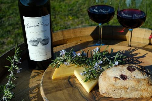 A bottle of red wine, two glasses, cheese, and bread on a wooden platter, surrounded by fresh herbs.