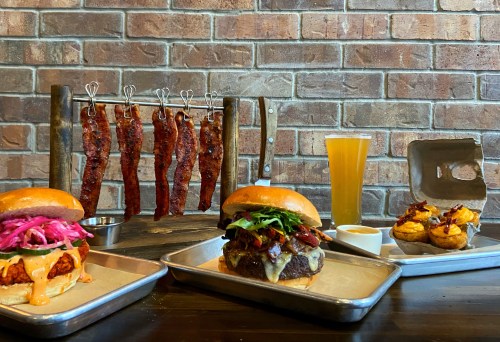A table with two burgers, a drink, and hanging strips of meat against a brick wall.
