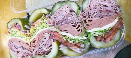 A container filled with stacked deli meats, lettuce, tomatoes, and pickles, arranged in a colorful display.