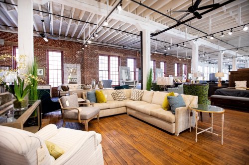 A spacious furniture showroom featuring a sectional sofa, armchairs, and decorative plants against a brick wall.