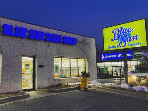 Exterior of Blue Sun Soda Shop at dusk, featuring bright signage and a nearby Sherwin-Williams store.