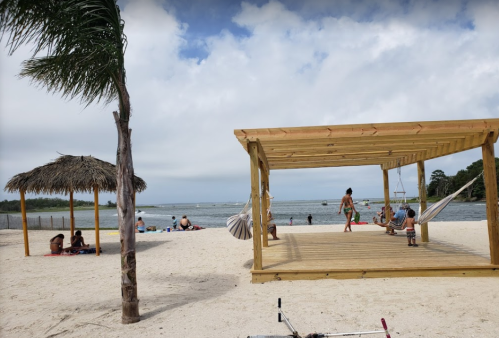 A sandy beach scene with a wooden pavilion, hammocks, and people relaxing under palm trees by the water.