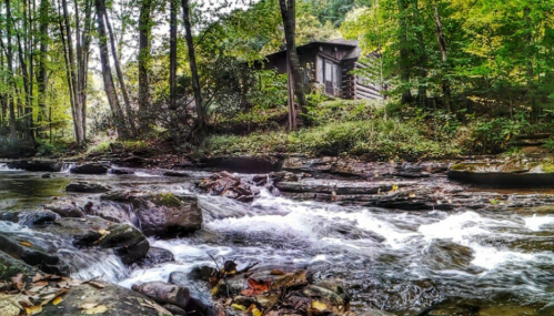 A serene stream flows past a log cabin, surrounded by lush green trees and autumn foliage.